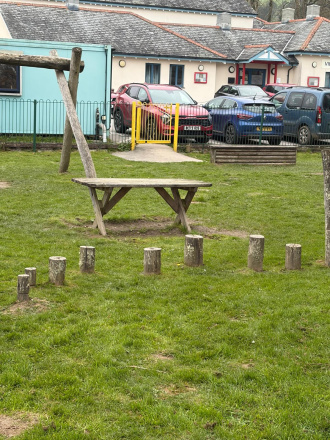 A wooden table in the playground. The village hall and several cars in the car park are behind.