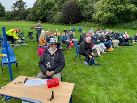 Simon Fann at a desk selling tickets on entry to the field. A grassy field behinf with audience in seats.