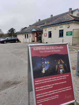 An A-frame sign advertising the 'caravan of stories' in the car park. The village hall is visible behind.